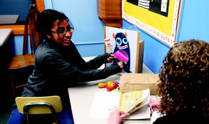 A smiling woman uses a colorful monster puppet during an activity with another person seated at a table with a picture book and toy food.
