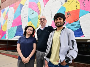 Three people stand smiling in front of a colorful mural featuring abstract portraits on a brick wall, with the artwork filling the background of the urban streetscape.