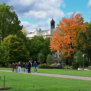 Visitors tour The College of Wooster campus along a walkway framed by green and orange fall foliage, with a historic stone building in the background.