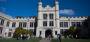 Kauke Hall at The College of Wooster, a light brick building with tall windows, turrets, battlements, and a central clock above an arched entryway. Students gather on the lawn and near the entrance, enjoying the sunny day under a bright blue sky.