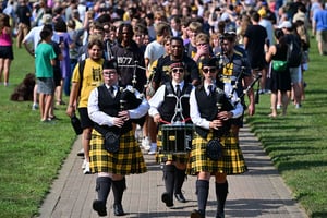 Three bagpipers and a drummer in black vests and yellow tartan kilts lead a large crowd of students walking along a campus path during a sunny outdoor event.