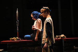 Two students in period costumes perform on a dimly lit theater stage beside a long table decorated with grapes and autumn leaves, with one kneeling in reflection and the other walking past wearing a floral wreath.